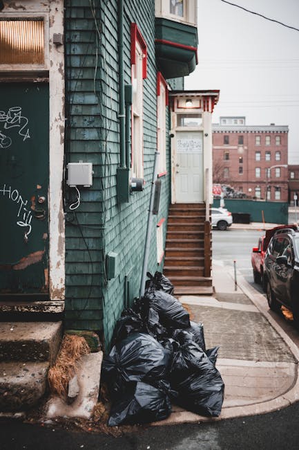 A row of black plastic rubbish bags piled against the side of a building with green wooden cladding and red-trimmed windows. The bags are stacked on a concrete sidewalk next to a small patch of frozen ground with patches of ice and snow. To the left, there is a graffiti-covered metal door with a small step in front, and an electrical box mounted on the wall above the bags. In the background, a set of wooden stairs with a handrail leads up to a white door at the building’s entrance, which has a small overhanging roof with red accents. Beyond the building, a street scene includes parked cars, a distant brick building, and overhead power lines. The overall environment suggests an area undergoing private rubbish collection or rubbish removal services, typical of a Bermondsey street or similar urban setting, with an emphasis on independent waste management practices.