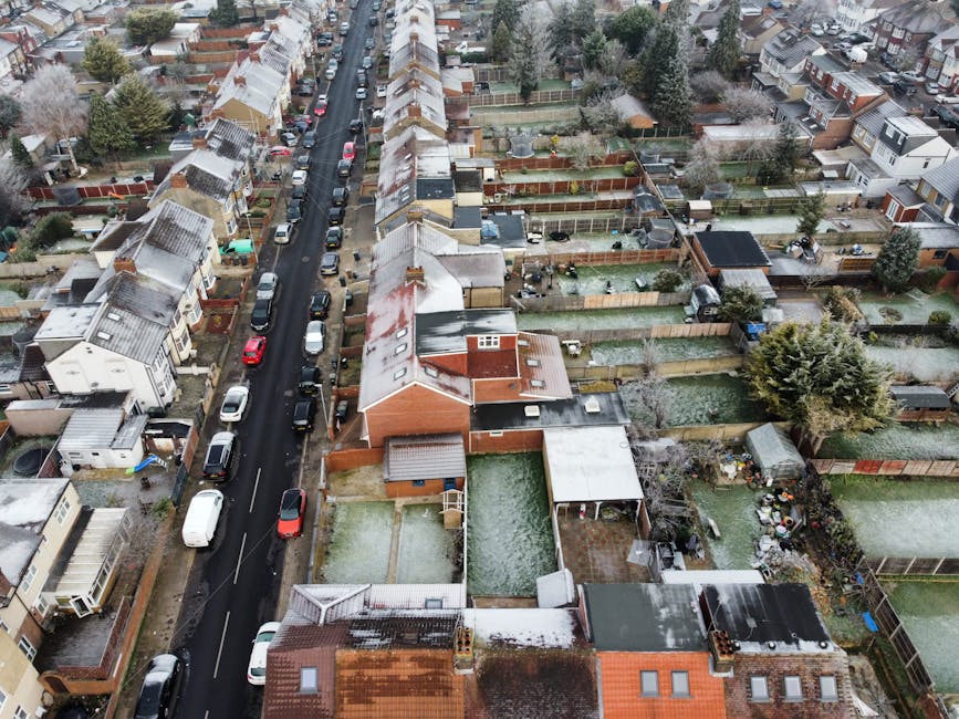 An aerial view of a residential neighborhood showing a narrow, straight street lined with parked cars on both sides. On the left side of the road, there are terraced houses with pitched roofs, some with snow or frost on their tiles, and small front gardens. To the right, there are back gardens enclosed by wooden fences, some with sheds, garden furniture, and neatly maintained lawns, all also dusted with light snow. The street appears quiet with a mix of personal vehicles, including hatchbacks, sedans, and vans, parked along the curb. Surrounding the houses are mature trees, some evergreen, providing greenery amidst the winter scene. The overall atmosphere is calm and typical of a suburban area during colder months. This type of environment often connects to private waste clearance services, such as those provided by Rubbish Clearance Bermondsey, supporting alternative rubbish removal and on-site clearance options for residents managing household or garden waste without reliance on local authority collections.
