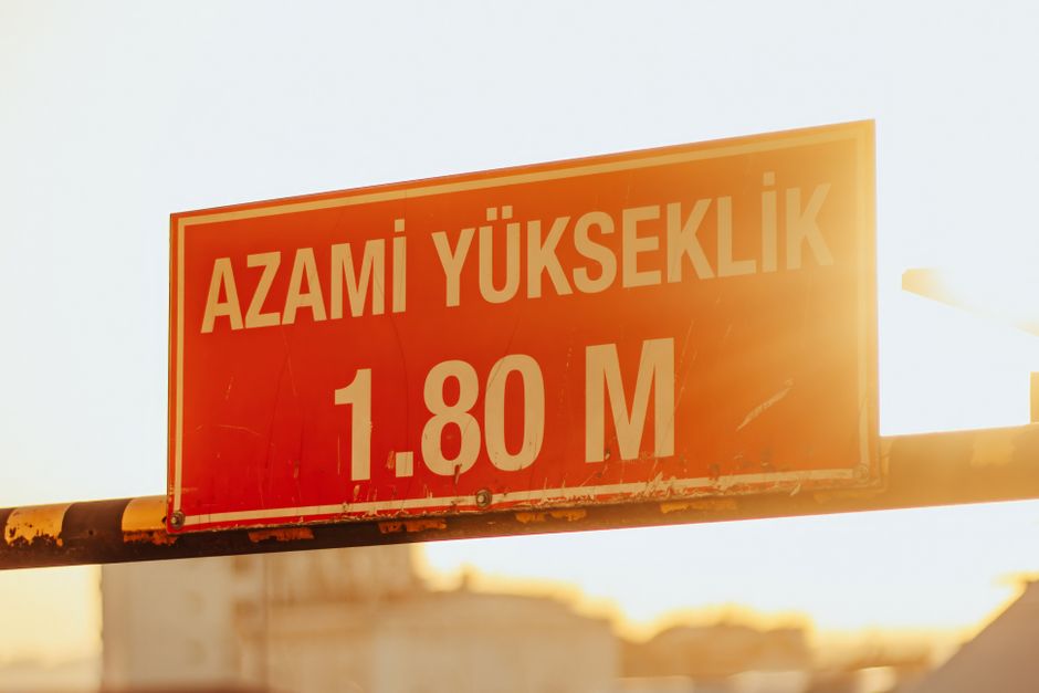 A rectangular orange and white road sign mounted on a rusty metal pole, displaying the message 'AZAMI YÜKSEKLİK 1.80 M' in bold capital letters, indicating a maximum height clearance of 1.80 meters. The sign is positioned outdoors against a bright, slightly cloudy sky, with the sunlight creating a warm glow on the sign's surface. In the background, out-of-focus building rooftops are visible, suggesting an urban setting where private waste or rubbish clearance services, such as those offered by Rubbish Clearance Bermondsey, might be relevant for managing bulky waste removal or on-site clearance tasks in the vicinity.