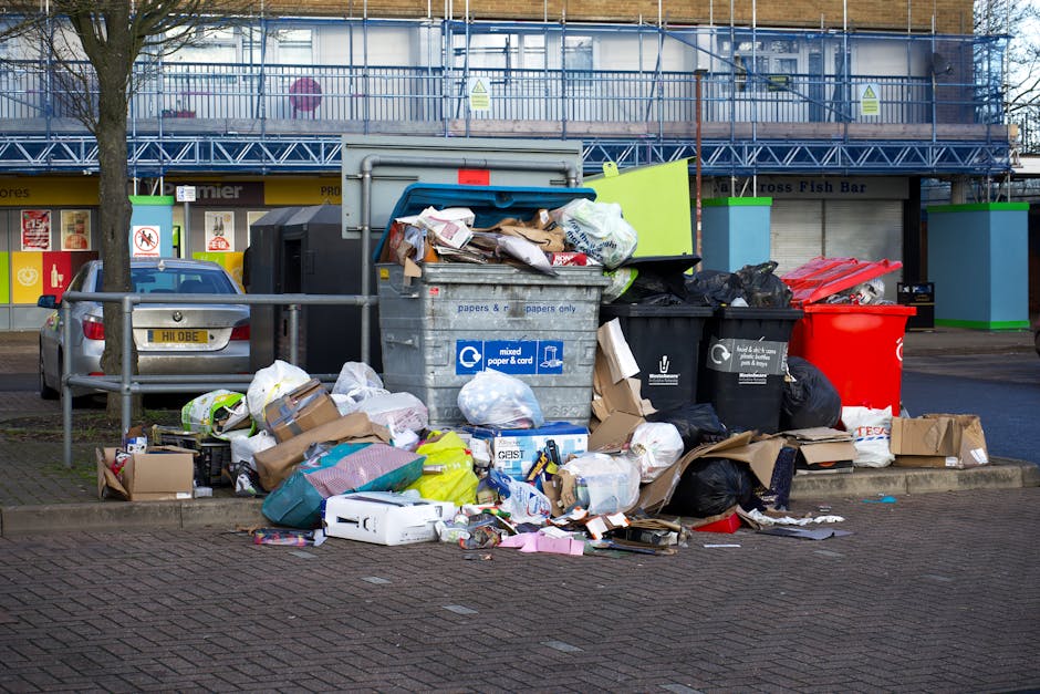 A large collection of mixed household waste and rubbish bags piled around and overflowing from a central grey recycling bin labeled for paper and cardboard, situated on a paved sidewalk in a commercial or urban area. Surrounding the bin are various discarded items including flattened cardboard boxes, plastic bags, crumpled paper, and loose packaging materials. To the left, parked behind a metal railing, is a small silver car with a visible yellow vehicle registration plate. In the background, there are storefronts with colorful signage and a building under renovation covered in blue scaffolding and protective netting. The scene is illuminated by natural daylight, suggesting a clear or partly cloudy day, with minimal shadows indicating diffused lighting. The overall setting reflects a typical public space where private rubbish collection or on-site clearance services like those offered by Rubbish Clearance Bermondsey might be utilized for alternative waste management outside council-provided waste disposal options.