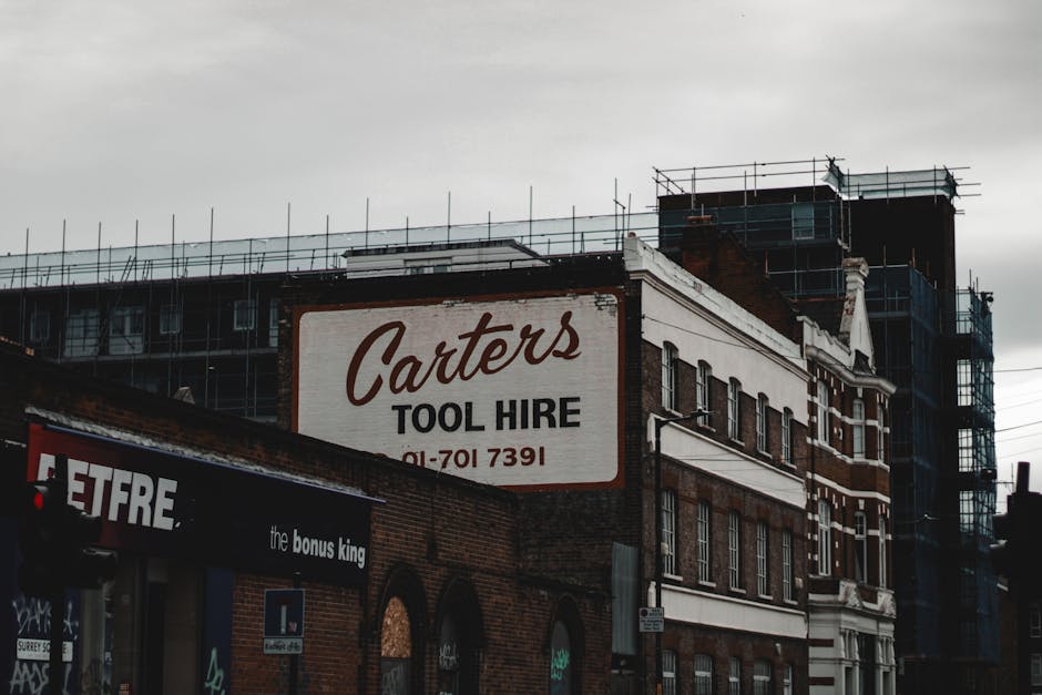 The image depicts a multi-story building with a partially finished upper section surrounded by scaffolding, indicating ongoing construction or renovation work. The foreground features a large, rectangular sign for 'Carter's Tool Hire' mounted on the side of the brick building, with a phone number underneath. Below this, part of another sign is visible, advertising 'the bonus king' store or business, with a portion of the store's signage on a black awning. The building’s exterior combines red brickwork with white decorative trim around the windows and along the upper edge. The sky above is overcast, casting diffuse light over the scene, which emphasizes the textured brick surfaces and the scaffolding. This scene exemplifies typical urban regeneration areas where buildings are being renovated or adapted, aligning with independent or private waste handling activities, such as rubbish removal or site clearance, often associated with building refurbishment projects. Rubbish Clearance Bermondsey would facilitate efficient emptying of construction debris from such sites, supporting alternative waste disposal solutions in busy city environments.
