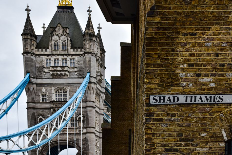 A close-up view of a brick wall with a street sign reading 'SHAD THAMES' attached to it, situated beside a narrow alleyway. In the background, part of the Tower Bridge in London is visible, with its iconic towers, intricately detailed masonry, and blue suspension cables stretching diagonally across the scene. The bridge's stonework includes gothic architectural elements and decorative turrets. The sky above is overcast, casting diffuse natural light over the scene, which emphasizes the texture of the weathered brick wall and the stone facade of the bridge. This urban setting highlights a typical view of the historic Tower Bridge juxtaposed with the modern street signage, relevant in the context of independent or private waste management services operating in the area, such as Rubbish Clearance Bermondsey, facilitating on-site waste removal or alternative rubbish disposal methods in central London.